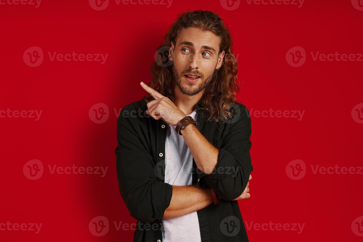 Good looking young man in casual clothing looking away and pointing copy space while standing against red background photo