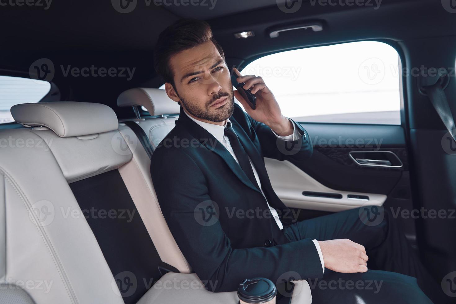 Día ocupado. un joven apuesto con traje completo hablando por teléfono inteligente y mirando la cámara mientras está sentado en el auto foto