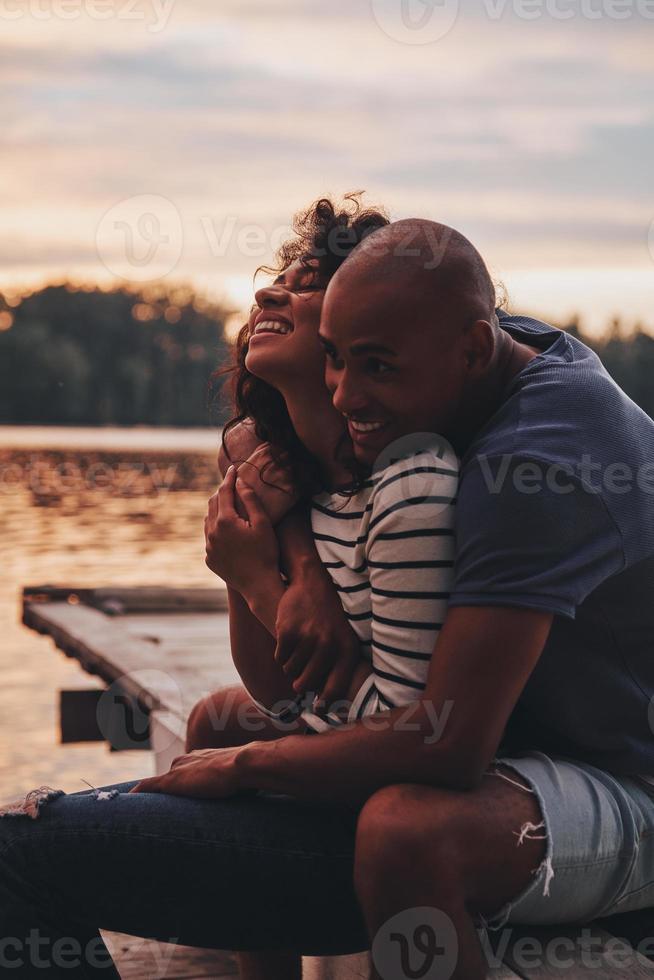 Too good to be true. Happy young couple embracing and smiling while sitting on the pier near the lake photo