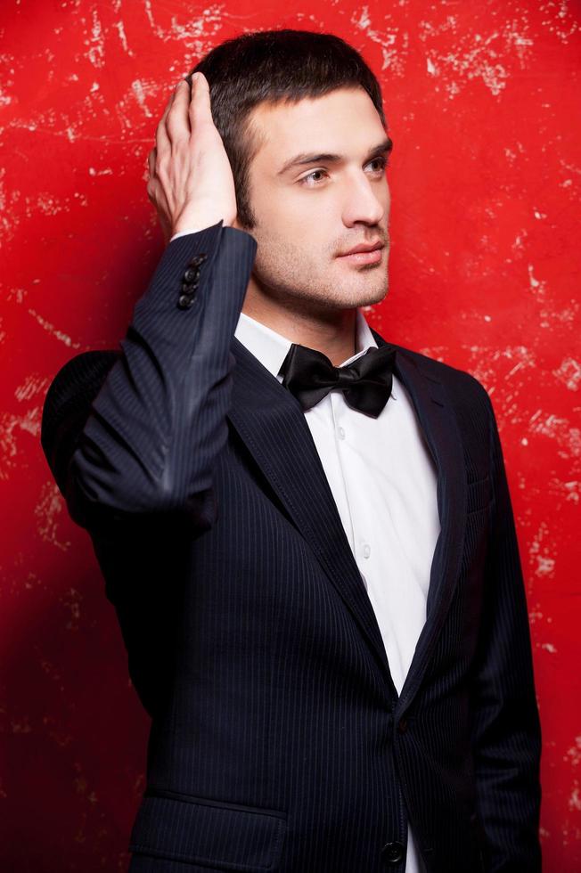 guapo y elegante. un joven apuesto con traje y corbata de moño tocándose el pelo y mirando hacia otro lado mientras se enfrenta a un fondo rojo foto