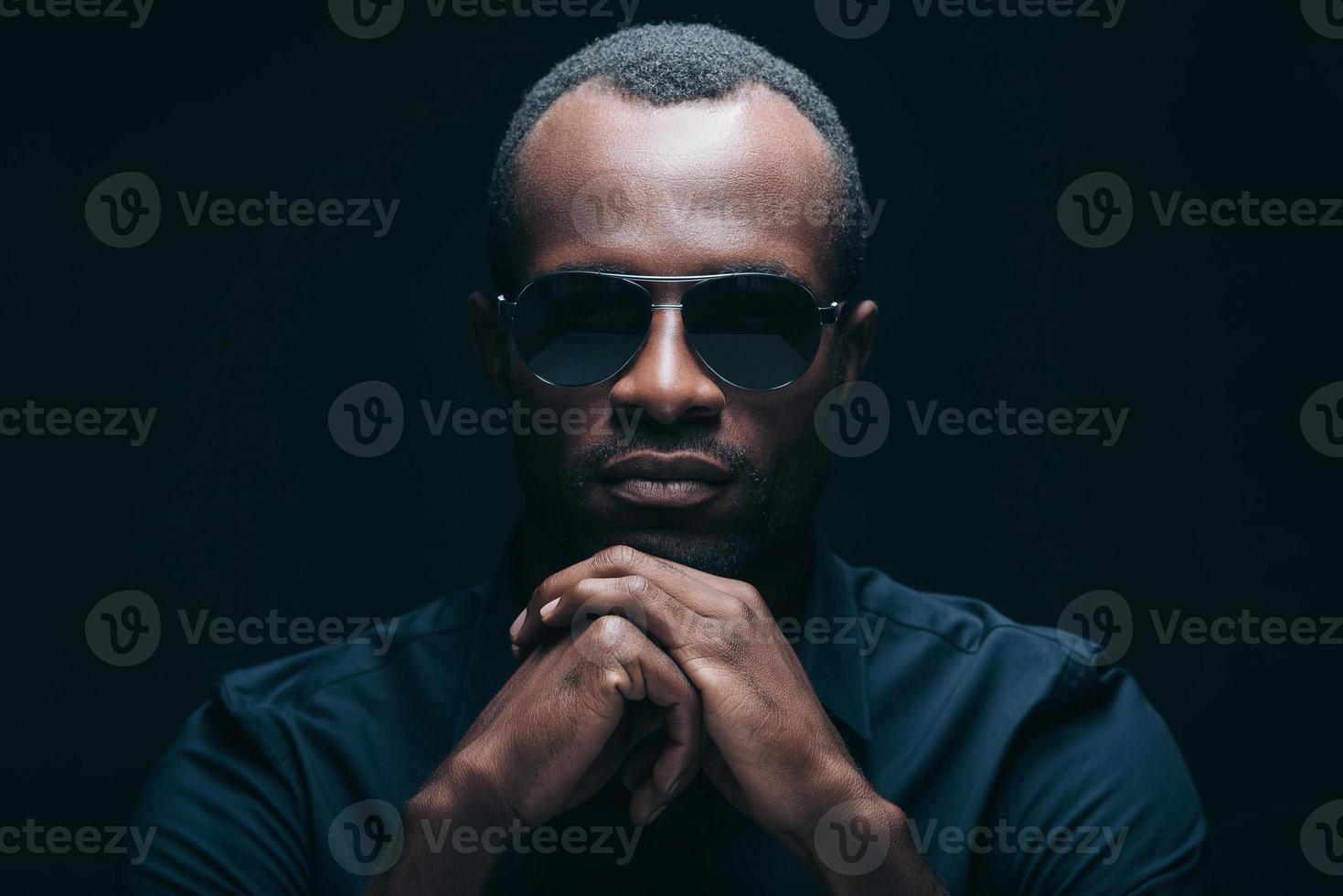 mirada segura y de moda. retrato de un apuesto joven africano con gafas de sol apoyando la cabeza en las manos y mirando a la cámara mientras está frente a un fondo negro foto