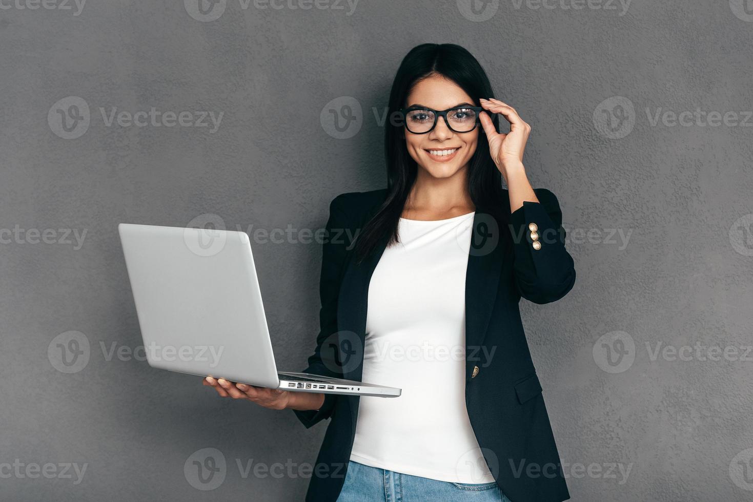 How may I help you Attractive young woman in smart casual wear and eyeglasses carrying laptop and smiling while standing against grey background photo