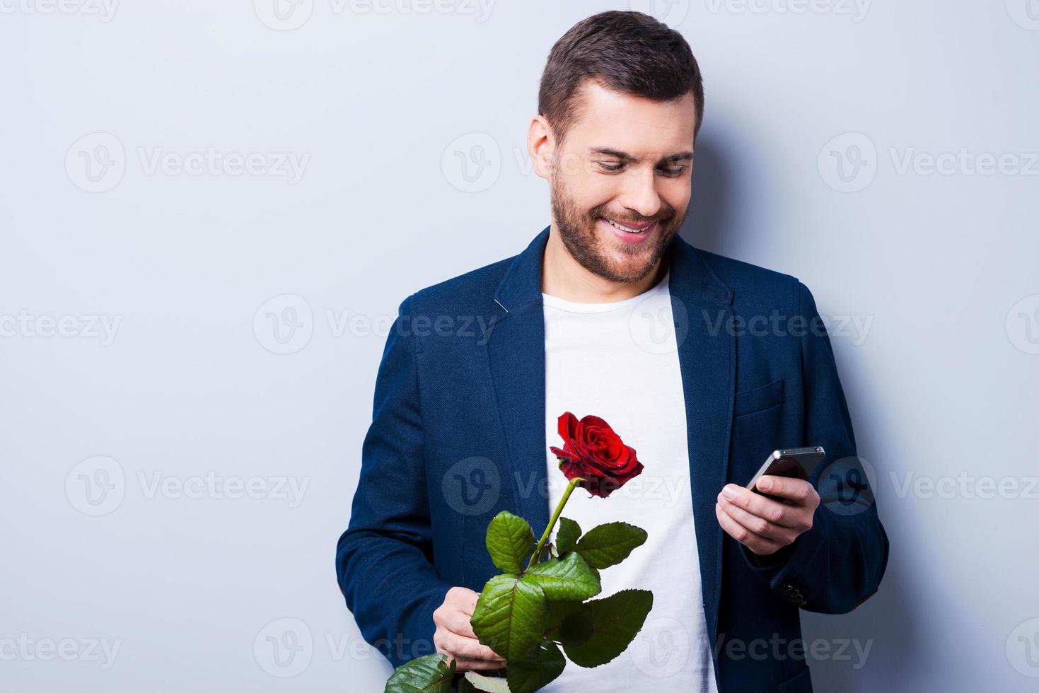 Waiting for her. Handsome young man typing message to his girlfriend while holding a flower and standing against grey background photo