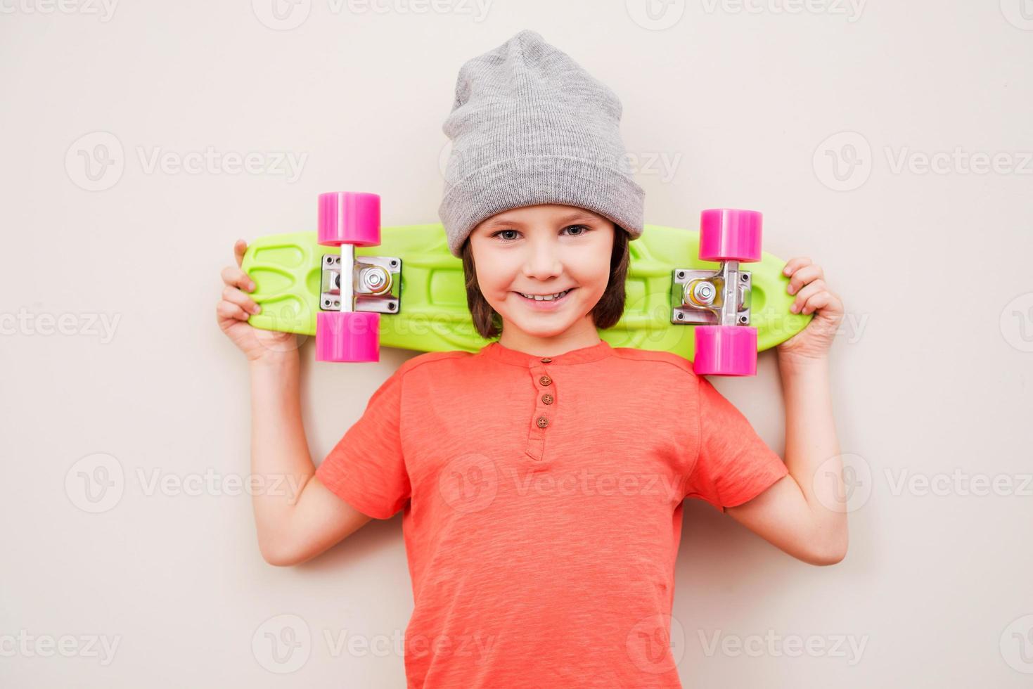 Ready to ride Happy little boy in hat holding colorful skateboard and smiling while standing against grey background photo