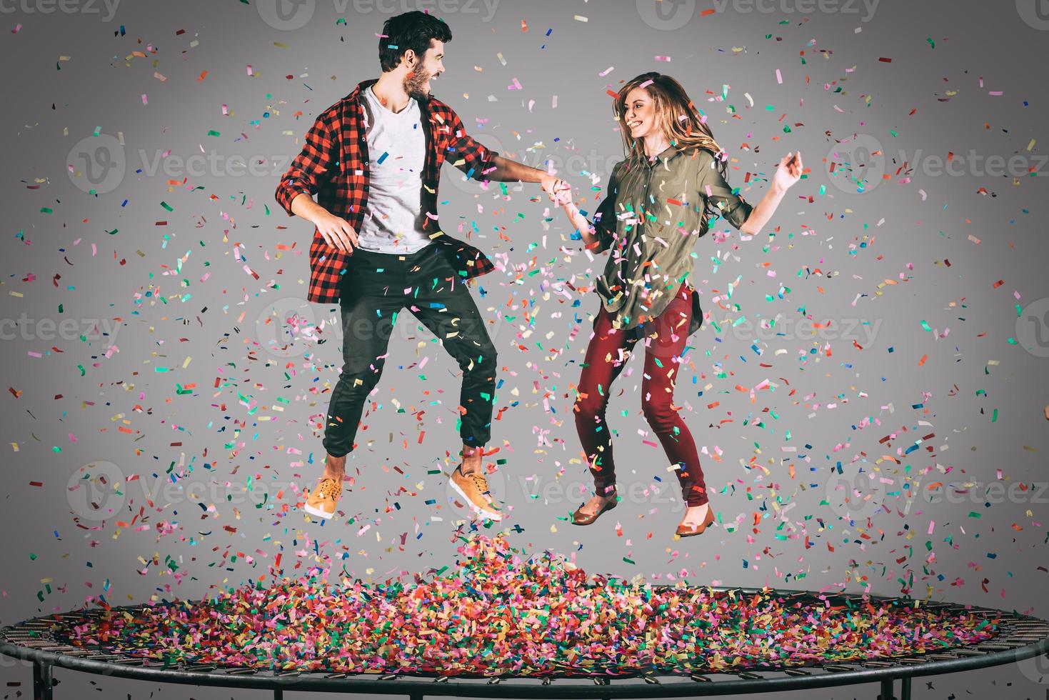 diversión colorida. toma en el aire de una hermosa pareja joven y alegre tomándose de la mano mientras salta en el trampolín junto con confeti a su alrededor foto