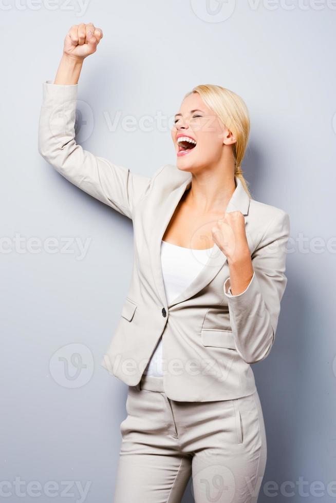qué día de suerte feliz joven empresaria manteniendo los brazos levantados mientras está de pie contra el fondo gris foto