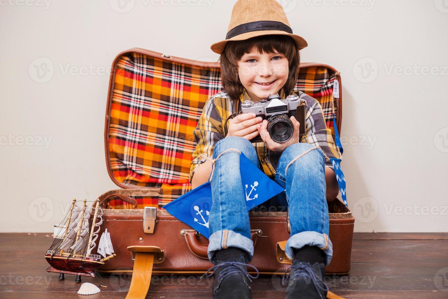 diga un niño de queso con sombreros sosteniendo una cámara y sonriendo mientras se sienta en una maleta con fondo marrón foto