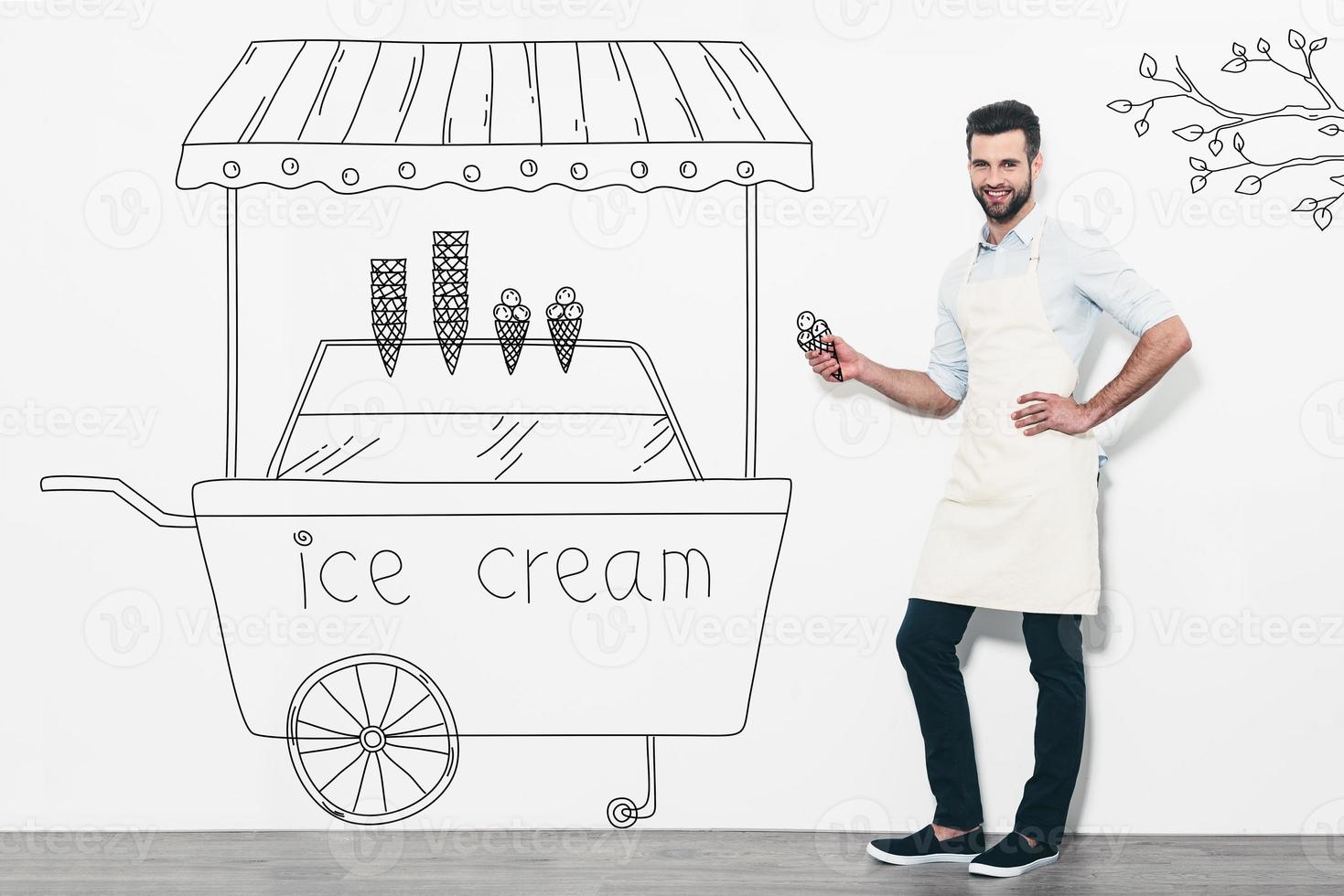 Selling the best ice cream. Handsome young smiling man in apron standing in front of the white wall and near the pencil drawn ice cream cart photo
