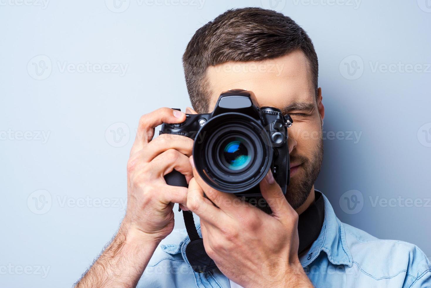 Focusing at you. Handsome young man photographing you while standing against grey background photo