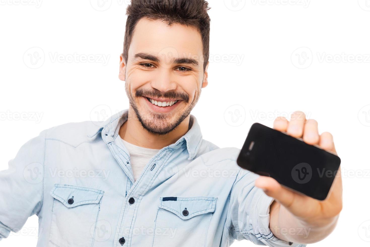 mira lo que tengo joven alegre que muestra el teléfono móvil y sonríe mientras está de pie contra el fondo blanco foto