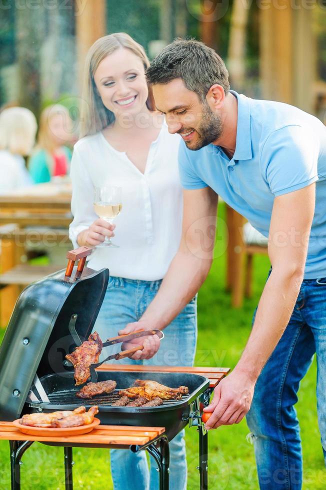 Need some help Happy young couple barbecuing meat on the grill while other members of family sitting at the dining table in the background photo