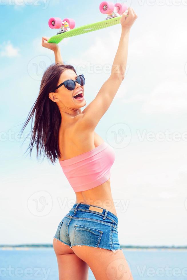 Ready to have fun Beautiful young woman holding colorful skateboard upon her head and smiling while standing at the riverside photo