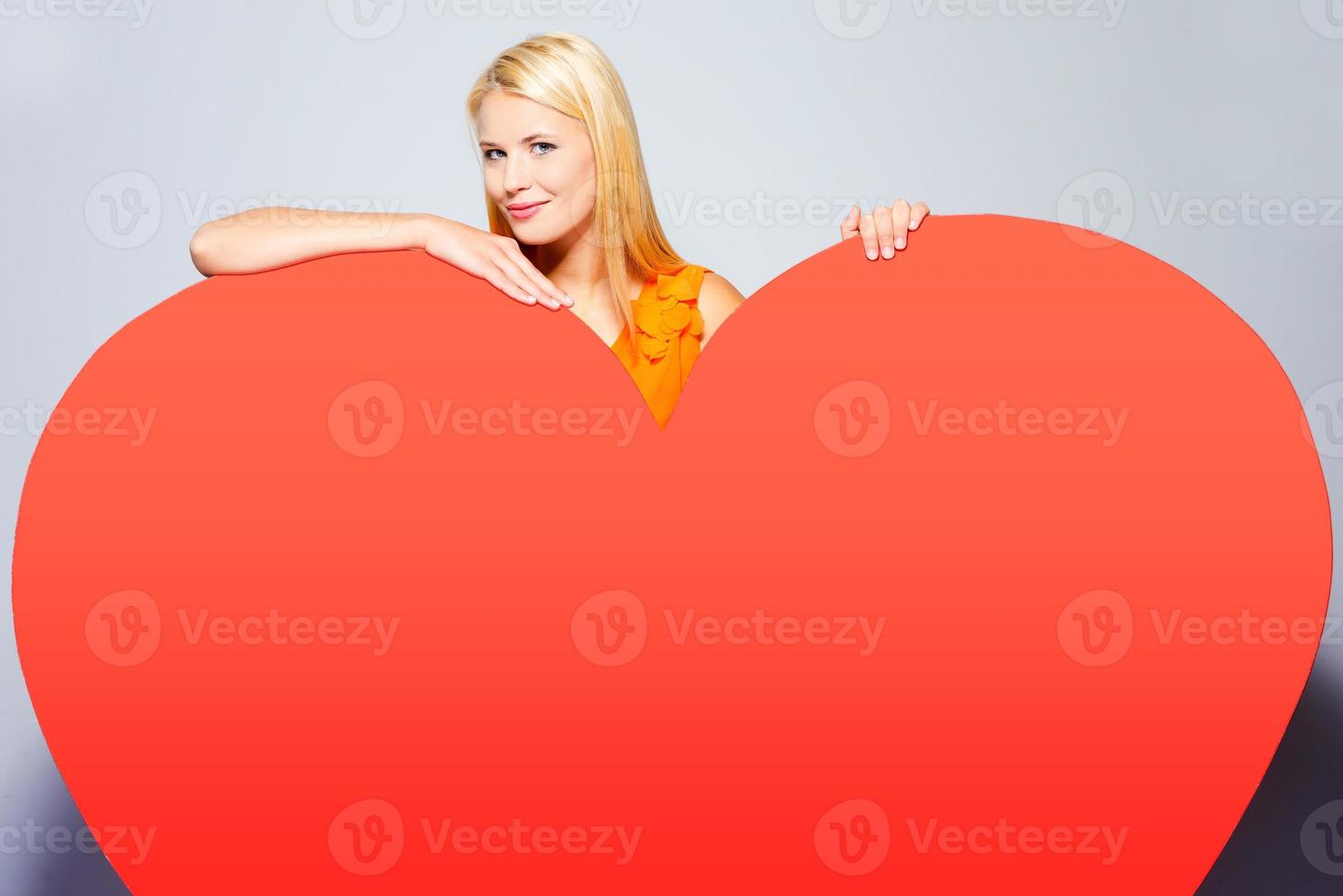 chica con gran corazón. hermosa joven rubia con un bonito vestido mirando a un enorme corazón rojo inclinado y sonriendo mientras se enfrenta a un fondo gris foto