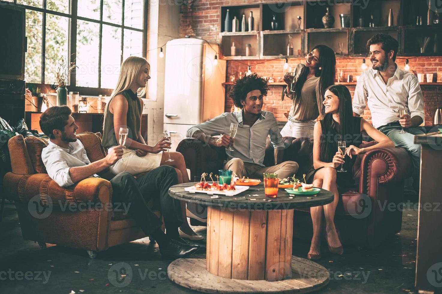 Friends will be friends. Group of cheerful young people enjoying food and drinks while spending nice time in cofortable chairs on the kitchen together photo