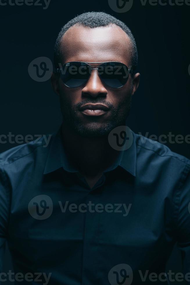 genial y guapo. retrato de un apuesto joven africano con gafas de sol mirando a la cámara mientras está frente a un fondo negro foto