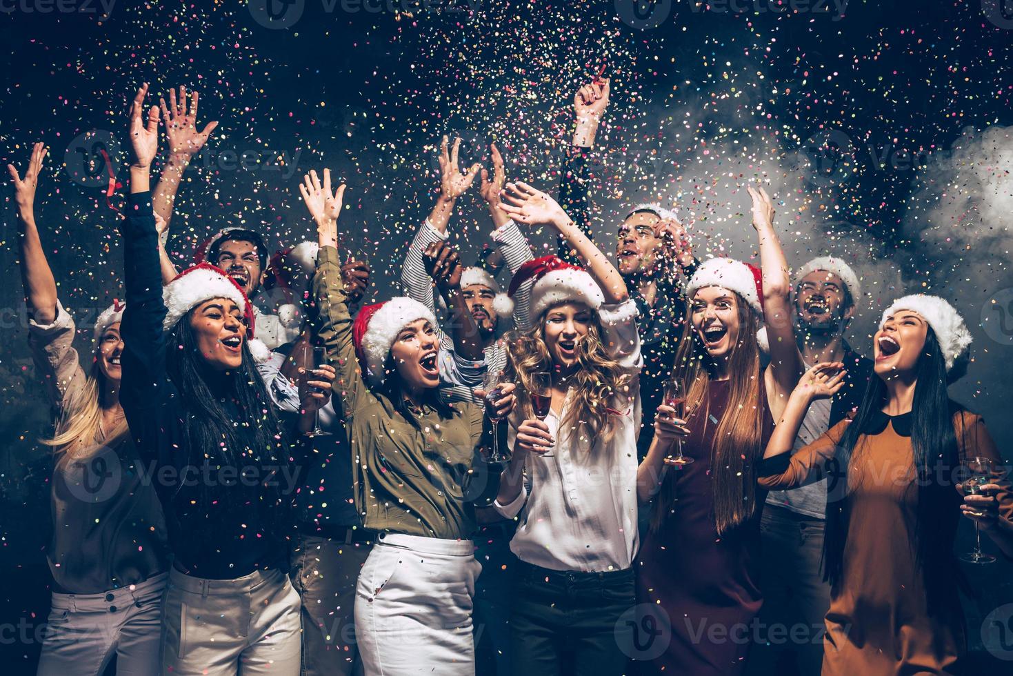 Celebrating New Year together. Group of beautiful young people in Santa hats throwing colorful confetti and looking happy photo