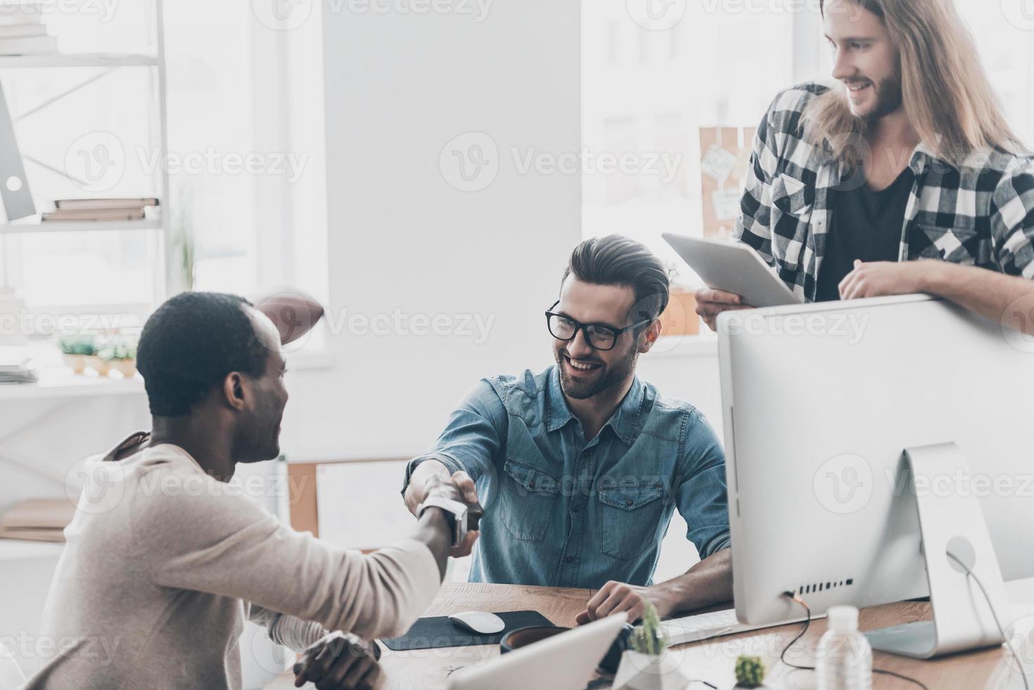 Welcome to our team Three businesspeople having a meeting while two of them shaking hands and smiling photo