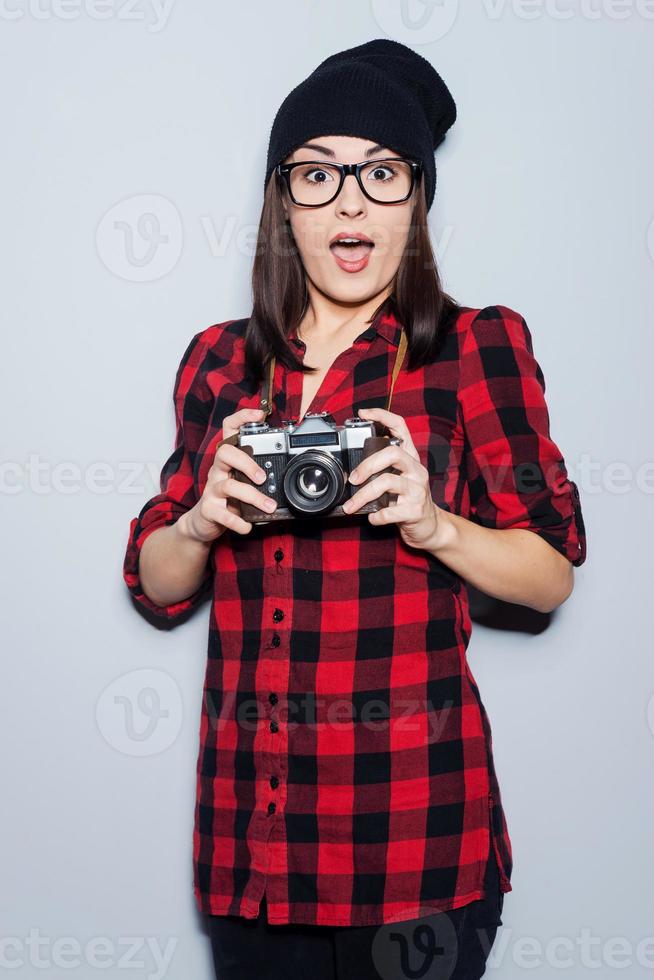 It can be a cool shot Surprised young woman in headwear and glasses holding camera and looking at camera while standing against grey background photo