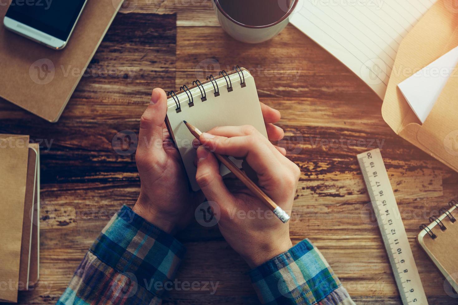 Making some urgent notes. Close-up top view of man holding notebook and writing something while sitting at wooden desk with different chancellery stuff laying on it photo