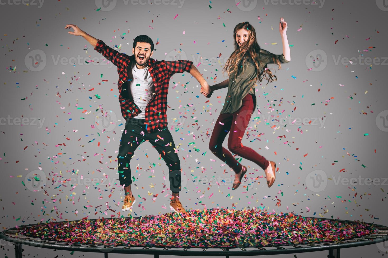 Fun in motion. Mid-air shot of beautiful young cheerful couple holding hands while jumping on trampoline together with confetti all around them photo