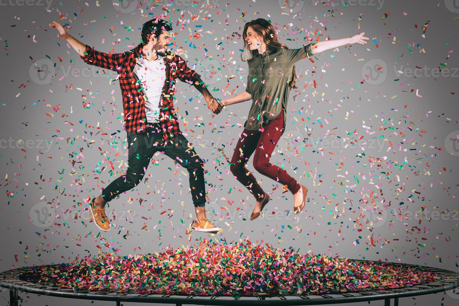 They love having fun. Mid-air shot of beautiful young cheerful couple holding hands while jumping on trampoline together with confetti all around them photo