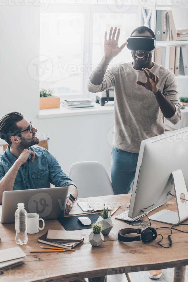 Enjoying new reality Two young successful men in casual wear working in creative office while one of them standing in VR headset photo