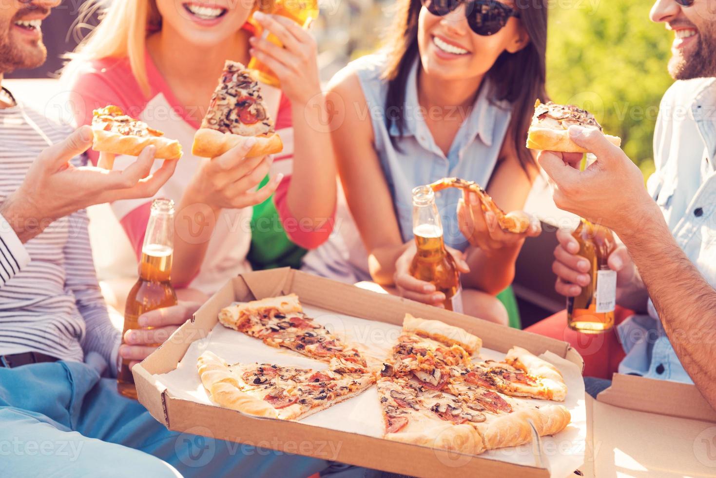 primer plano de la hora de la pizza de cuatro jóvenes alegres comiendo pizza y bebiendo cerveza al aire libre foto