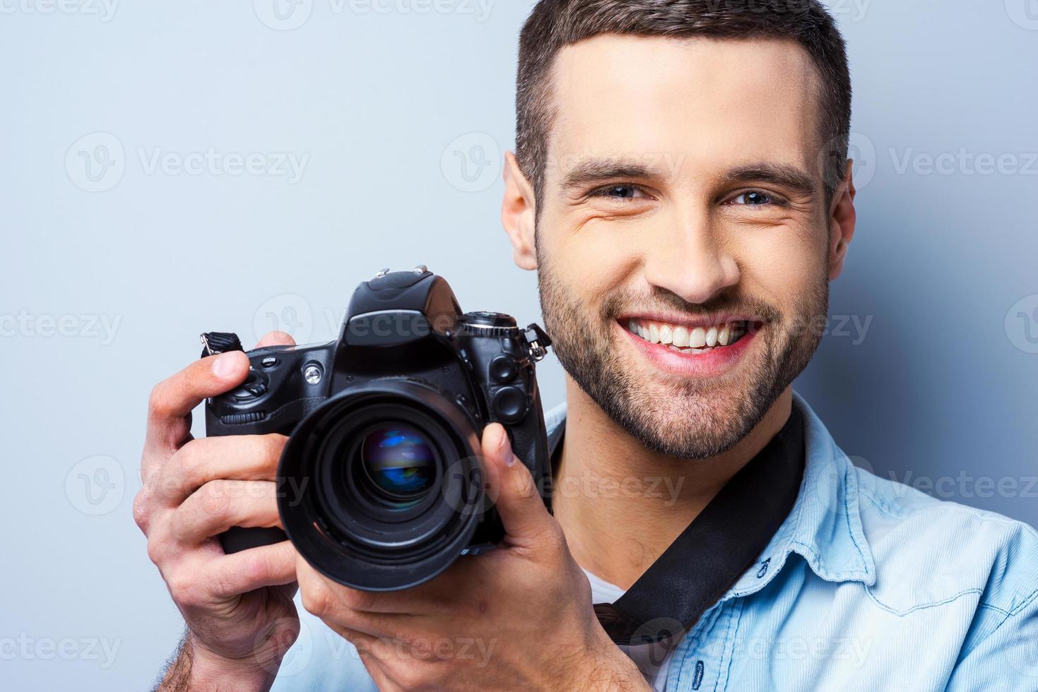 sonríe a una cámara un joven apuesto que sostiene una cámara digital y sonríe mientras se enfrenta a un fondo gris foto