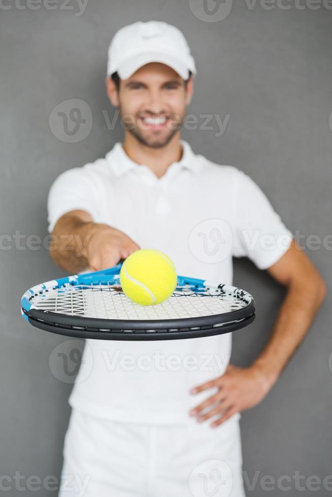 Play with me Happy young man in sports clothes stretching out tennis racket with ball and smiling while standing against grey background photo