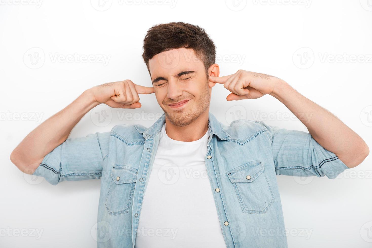 This is too loud Frustrated young man holding fingers in ears and keeping eyes closed while standing against white background photo
