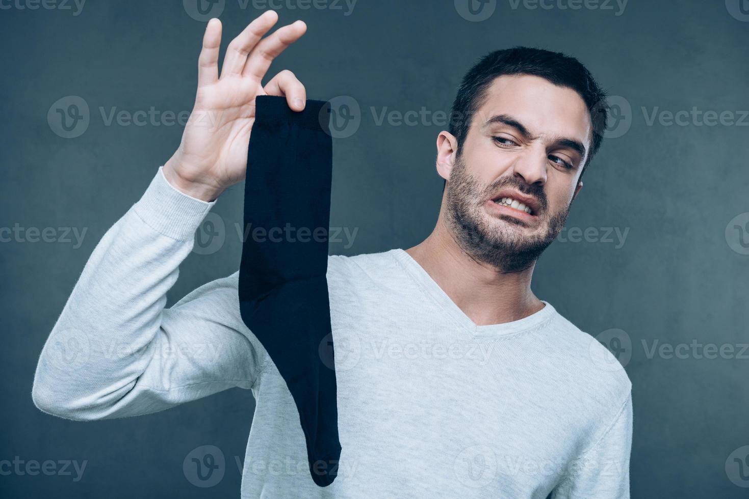 What is that Frustrated young man expressing negativity and covering nose with fingers while holding black sock and standing against grey background photo