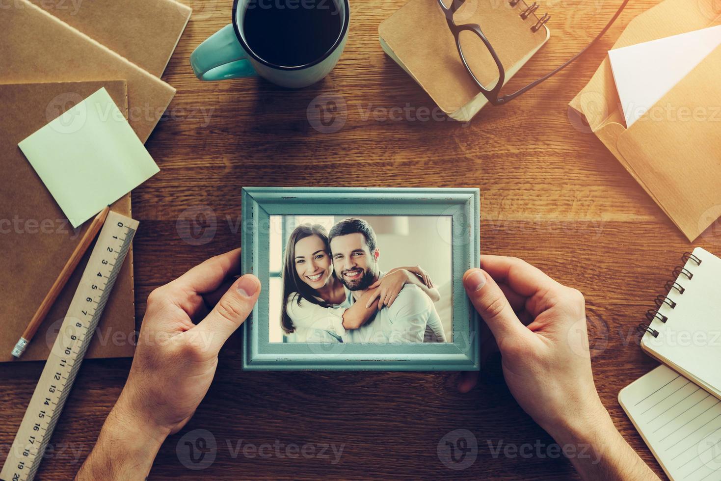Bright moments together. Close-up top view of man holding photograph of young couple over wooden desk with different chancellery stuff laying around photo