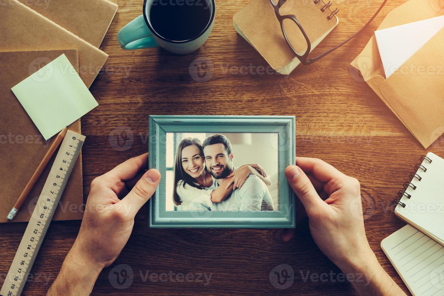 momentos brillantes juntos. vista superior de primer plano del hombre que sostiene la fotografía de una pareja joven sobre un escritorio de madera con diferentes cosas de la cancillería tiradas por ahí foto