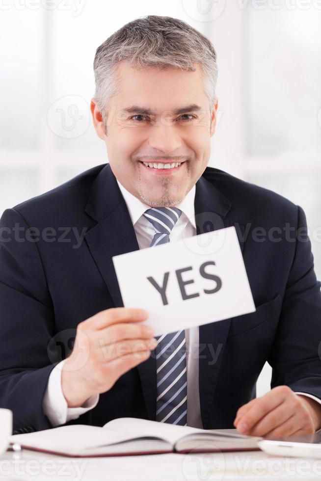 Approved Cheerful grey hair man in formalwear holding a paper with yes sign on it while sitting at her working place photo