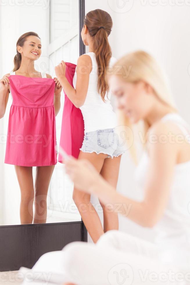 Maybe this dress Beautiful young woman holding dress and looking at the mirror while another women sitting on the foreground and looking at the mobile phone photo