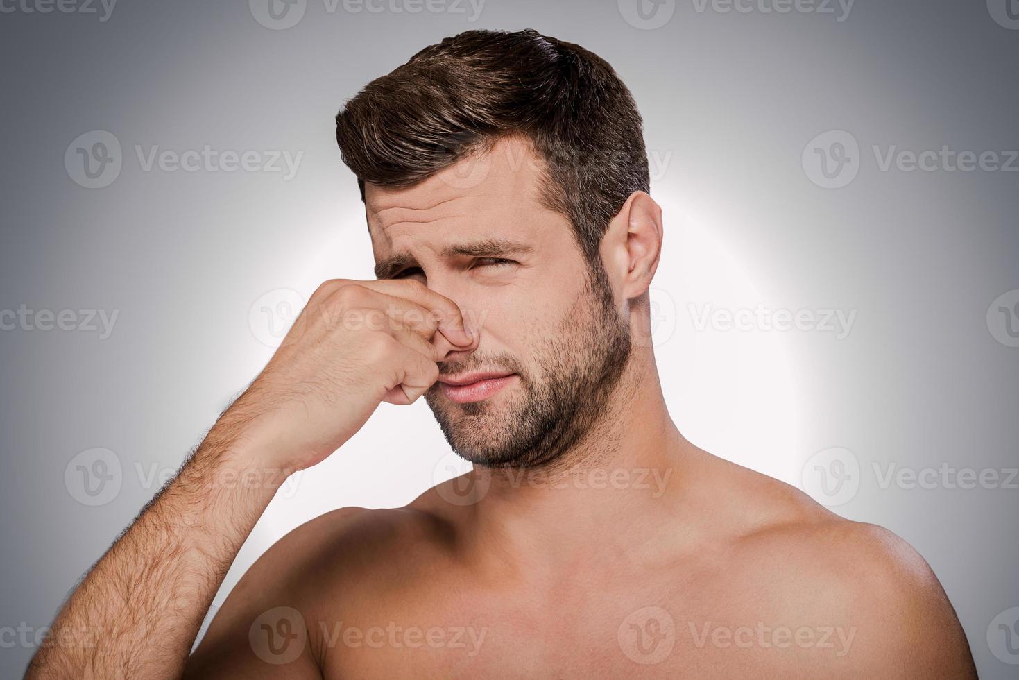 This awful smell Portrait of frustrated young shirtless man holding fingers on nose and making a face while standing against grey background photo