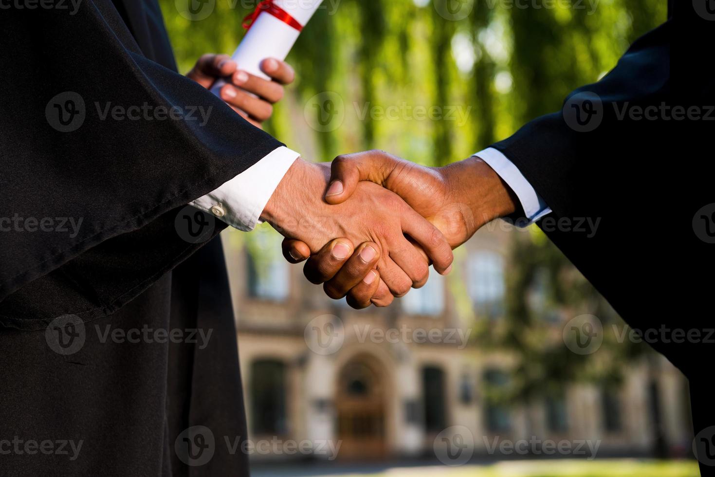 Congratulations Close-up of two men in graduation gowns holding diplomas and shaking hands photo