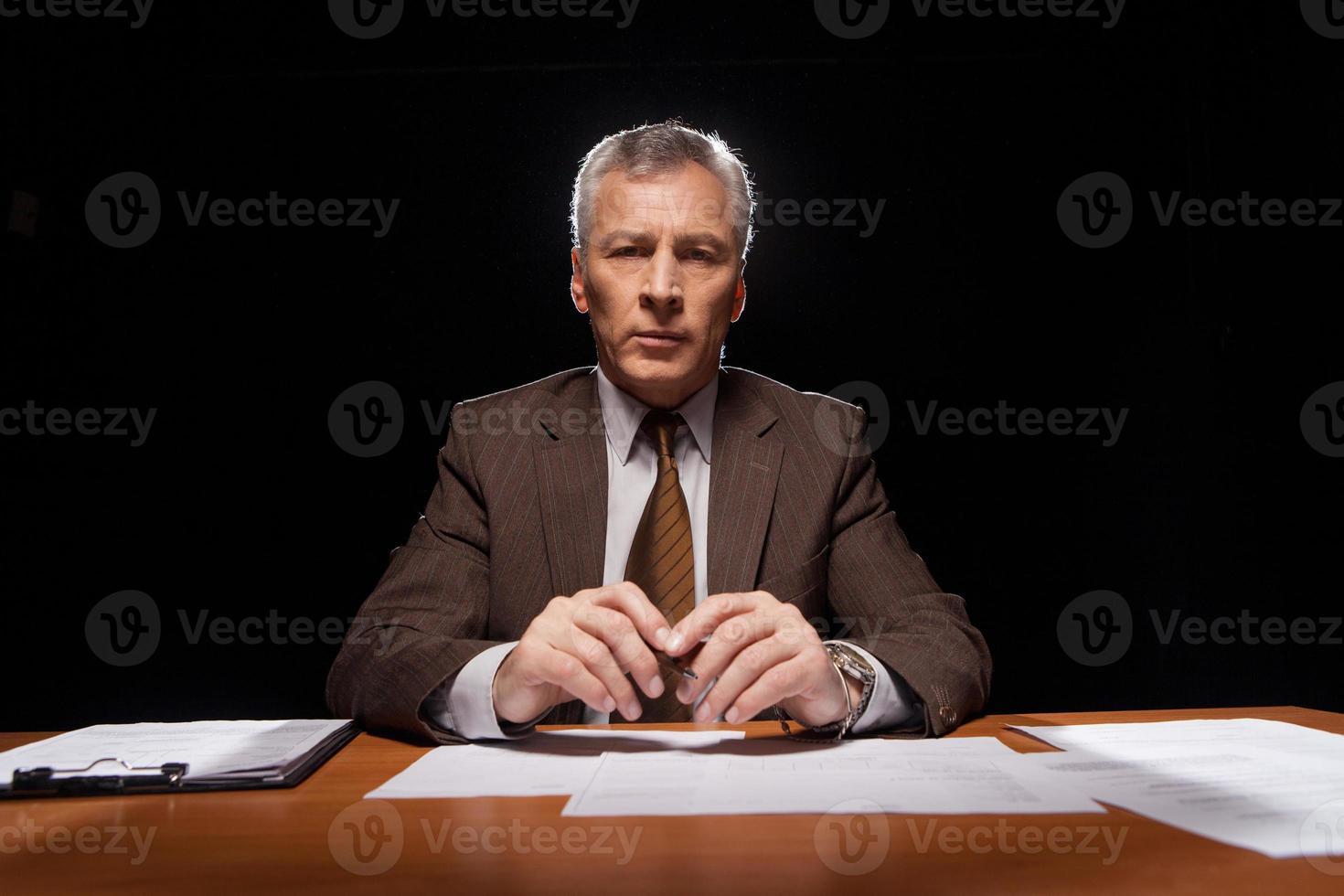 I am a boss Serious senior man in formalwear sitting at his working place and looking at camera while isolated on black background photo