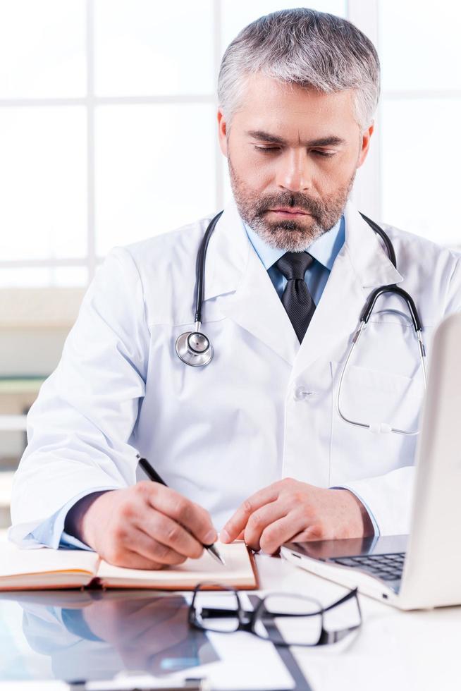 Confident doctor at work. Mature grey hair doctor writing something in note pad while sitting at his working place photo