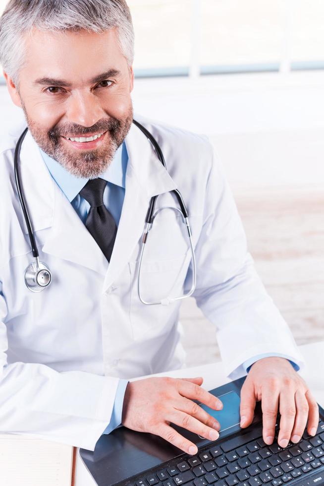 Confident and experienced doctor. Top view of confident mature grey hair doctor working on laptop and smiling while sitting at his working place photo