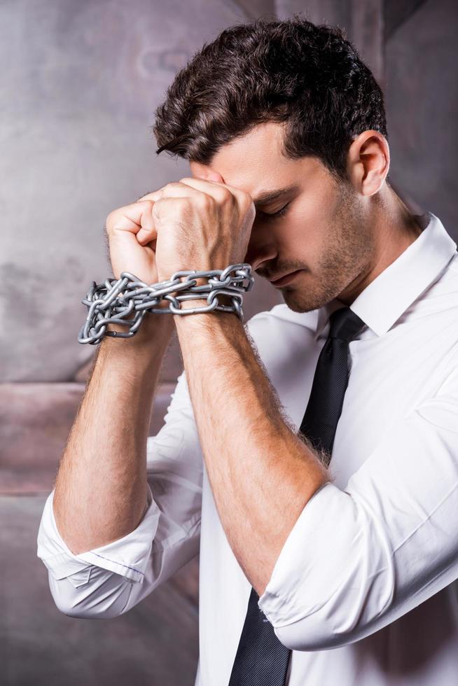 Trapped in chains. Side view of frustrated young man in shirt and tie touching his forehead with hands trapped in chains photo
