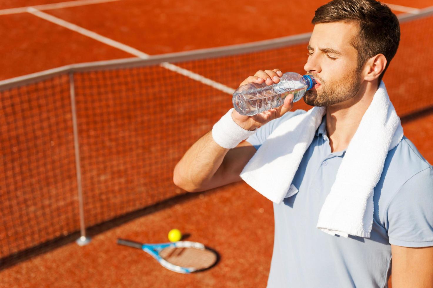 refrescarse después del juego. joven sediento con camisa de polo y toalla en los hombros bebiendo agua mientras está de pie en la cancha de tenis foto