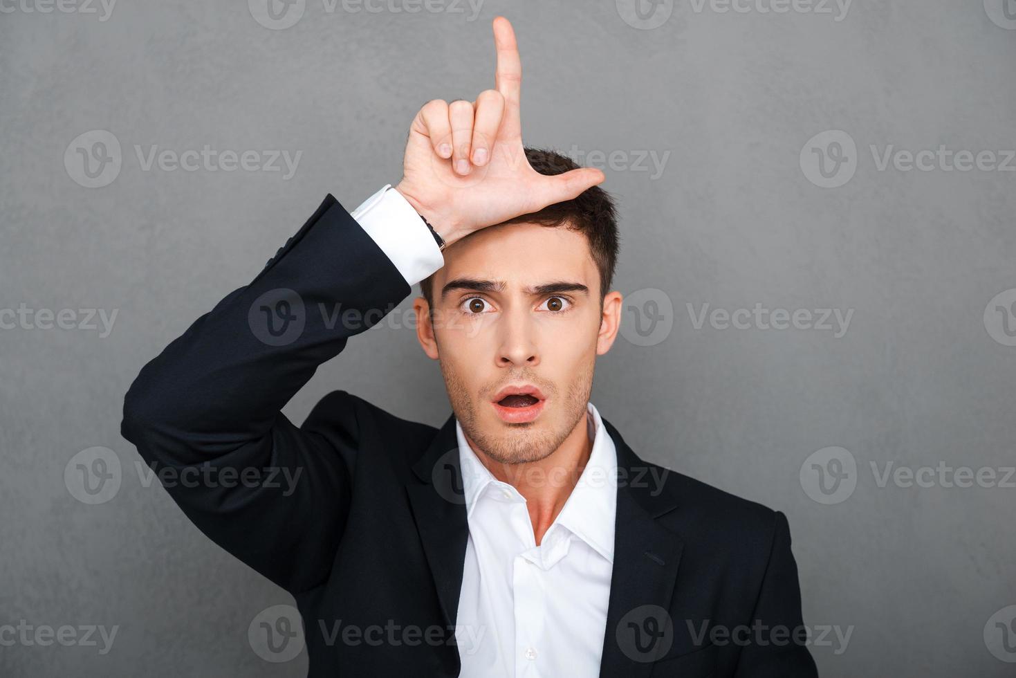 Loser Frustrated young man in formalwear gesturing loser sign and looking at camera while standing against grey background photo