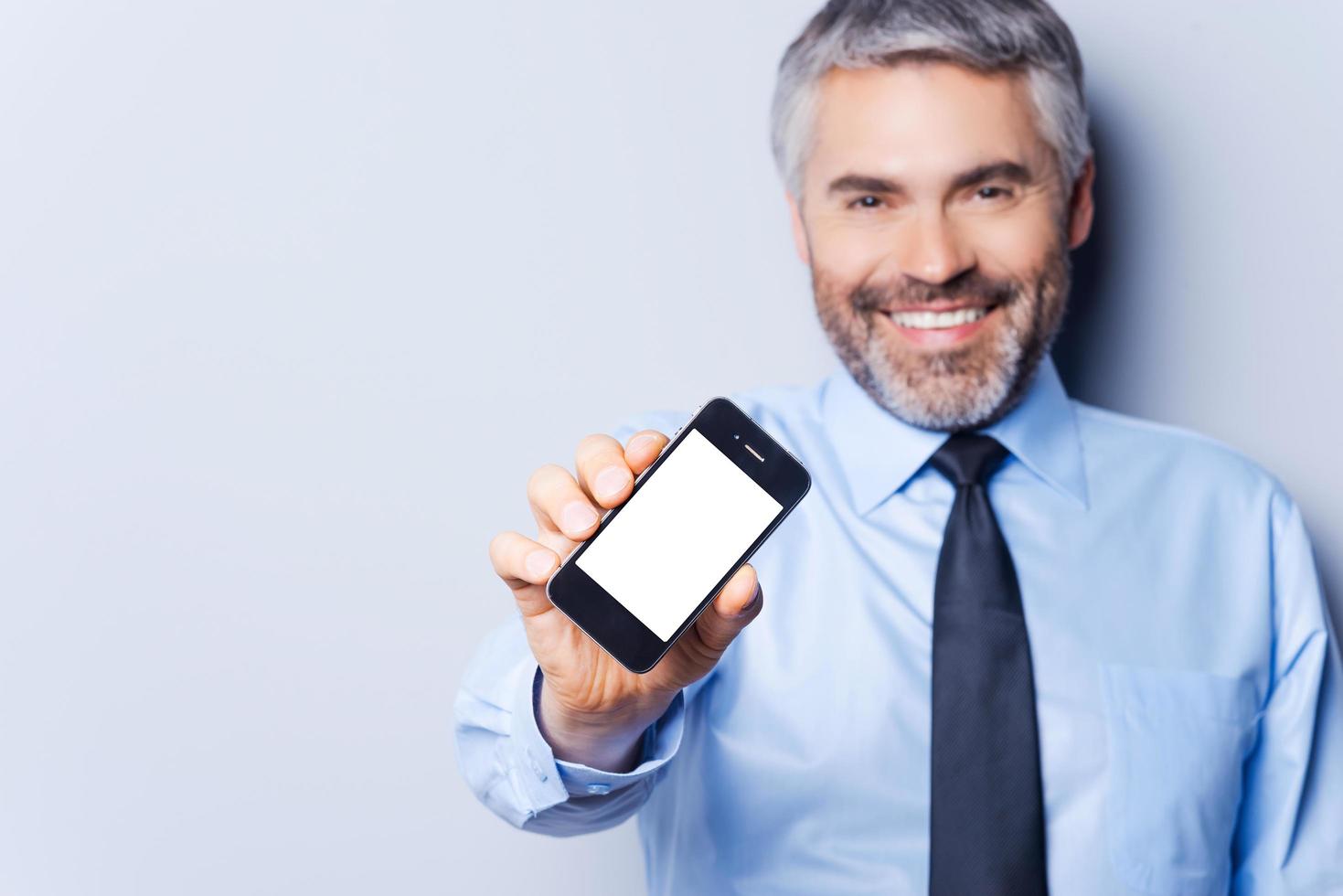 hombre de negocios feliz con teléfono móvil. hombre maduro feliz con camisa y corbata mostrando su teléfono móvil y sonriendo mientras se enfrenta a un fondo gris foto