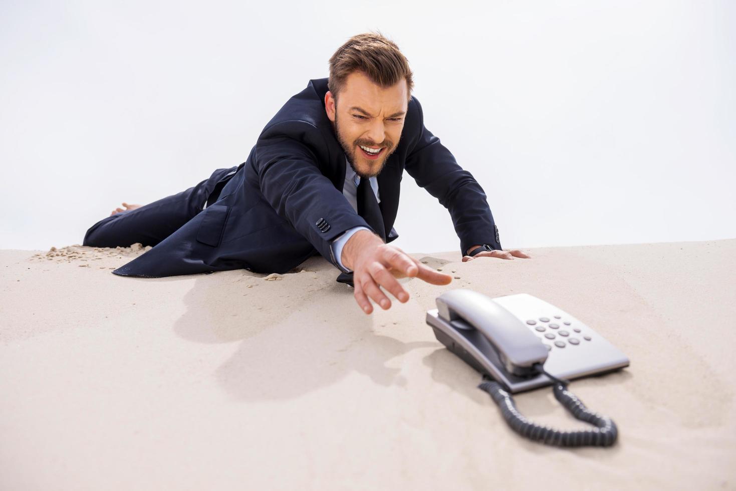 Urgent call. Young businessman stretching hand to telephone laying on sand photo