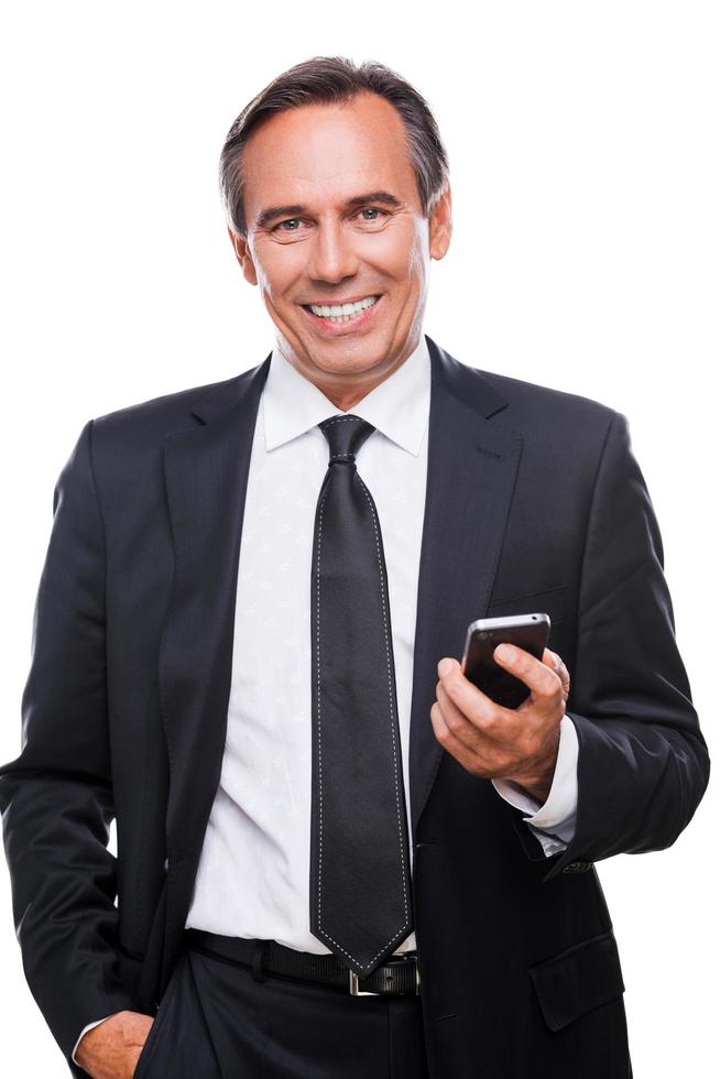 Always staying connected. Confident mature man in formalwear holding mobile phone and smiling while standing isolated on white background photo