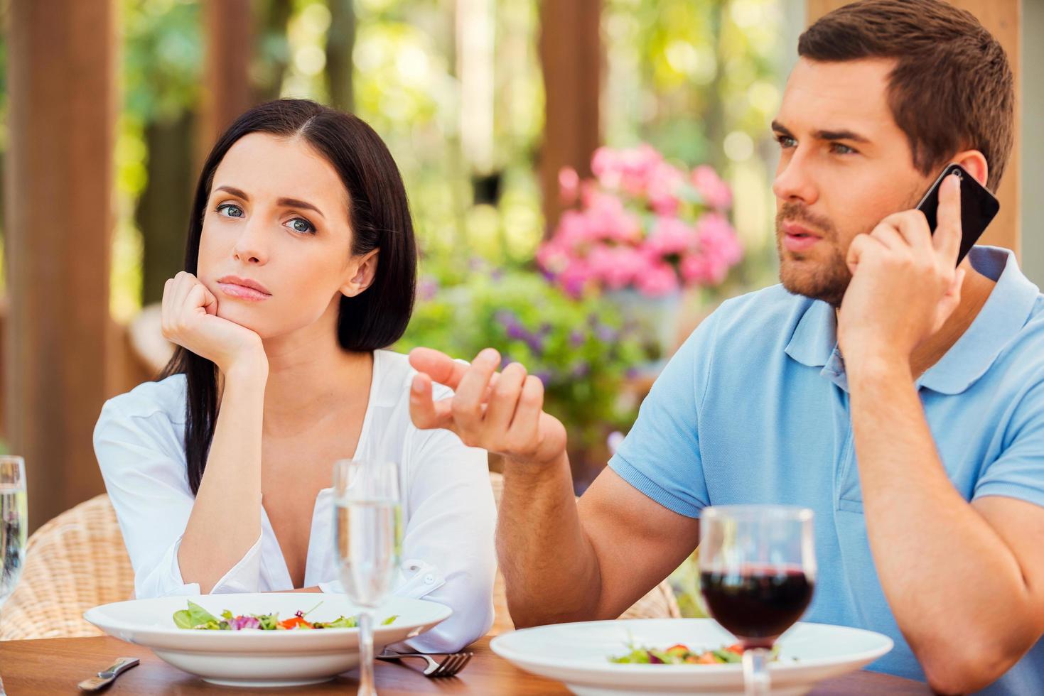 He always ignoring me. Depressed young woman holding hand on chin and looking at camera while her boyfriend talking on the mobile phone at outdoors restaurant photo