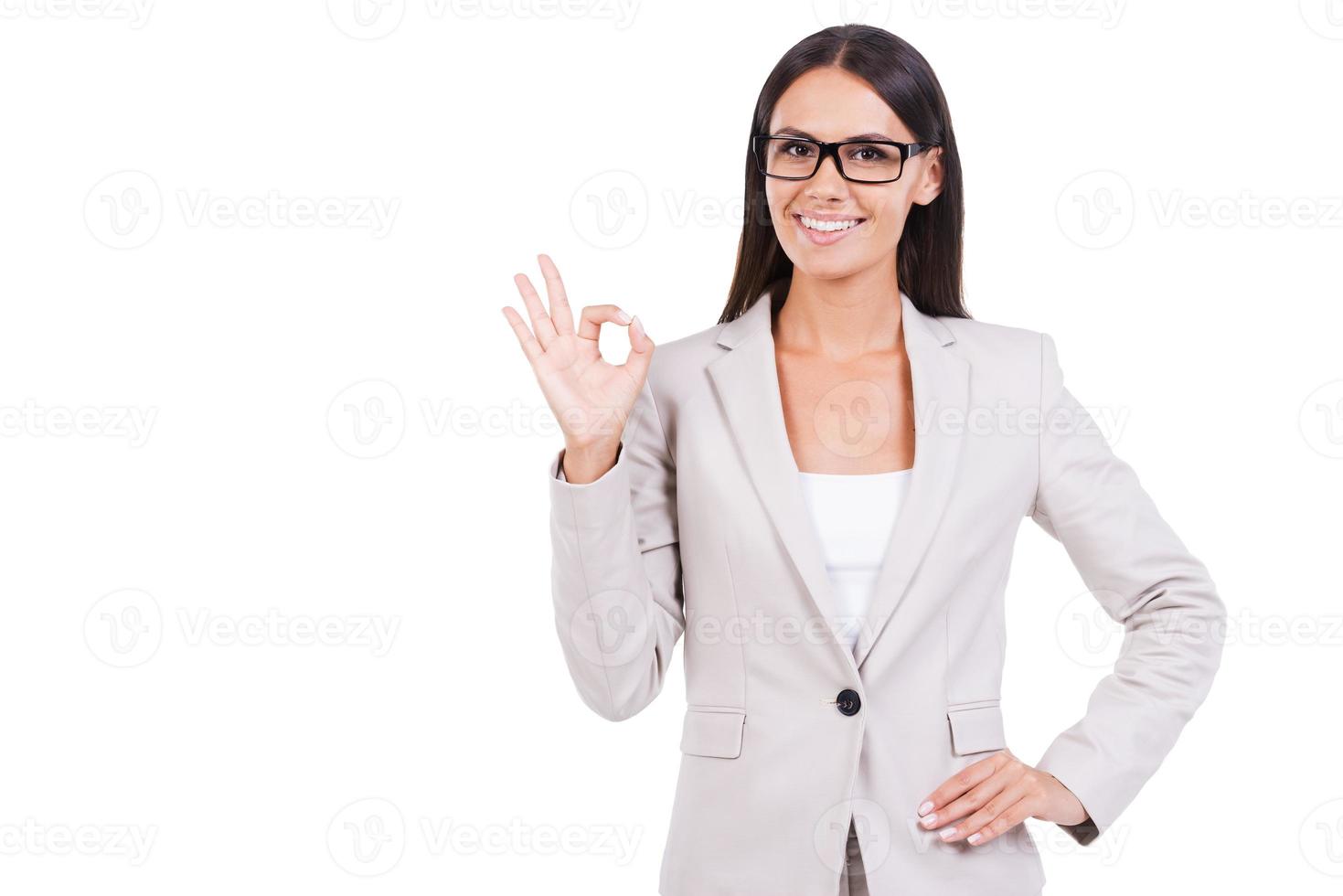 todo está bien. hermosa joven mujer de negocios en traje haciendo un gesto de ok y sonriendo mientras está de pie contra el fondo blanco foto