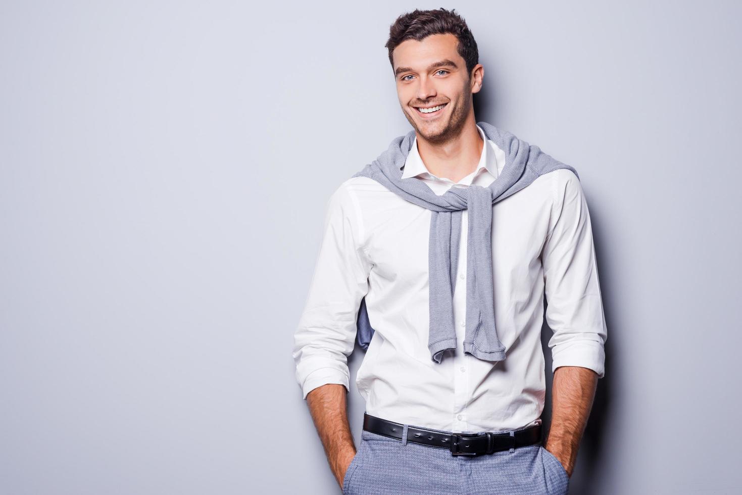 Handsome in style. Cheerful young man in smart casual wear looking at camera and holding hands in pockets while standing against grey background photo