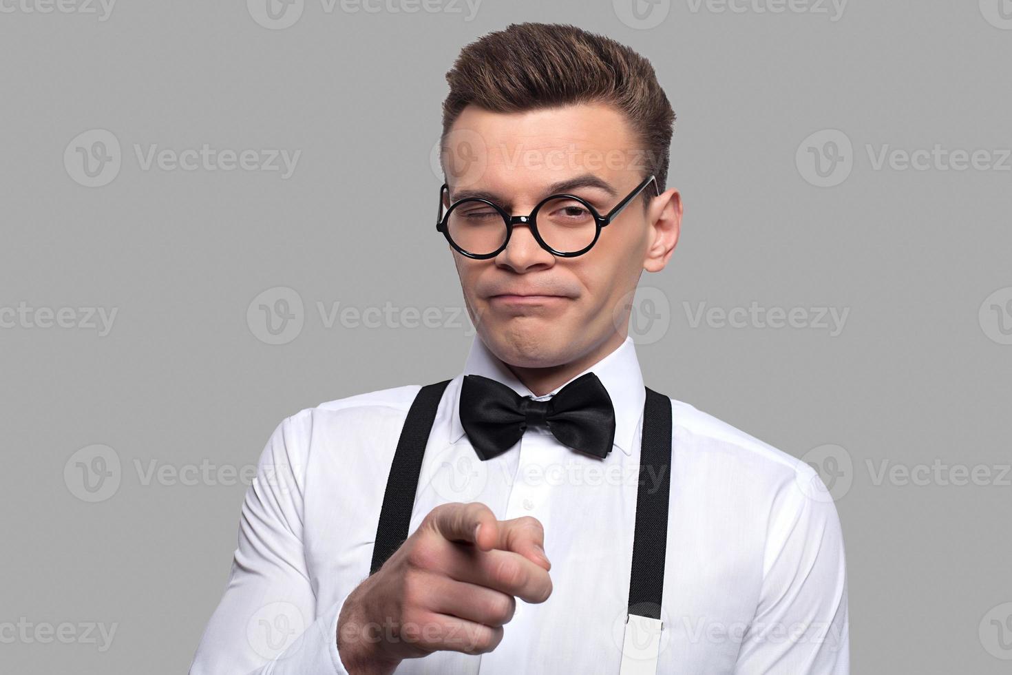 Elijo tu retrato de un joven nerd con corbata de moño y tirantes apuntando a la cámara mientras está de pie contra un fondo gris foto